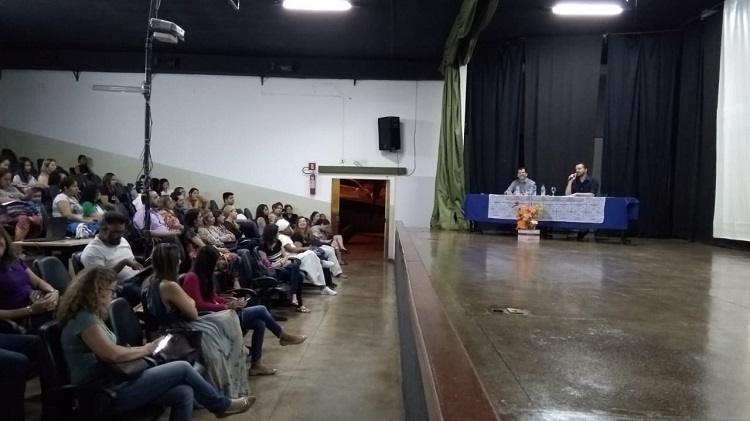 Mostra o palco e a plateia. No palco, há o palestrante e o coordenador do programa, sentados, atrás de uma mesa com forro azul e branco