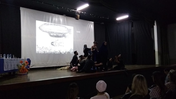 Foto escura com 10 atores, sendo 3 em pé, roupas escuras. No painel, um zepelim no céu e uma mulher deitada.