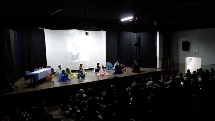 13 atores sentados no chão, cobertos, cada um por um lenço grande. Cada lenço de uma cor diferente.