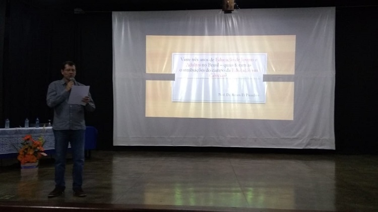 Professor Paulo Henrique, em pé, sobre o palco, lendo um papel que ele segura. Calça jeans e camisa cinza