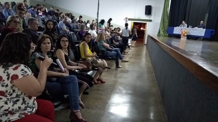 Professora Luciene, sentada na plateia, segurando um microfone, faz uma pergunta ao palestrante, que está sentado atrás de uma mesa no palco. Ela, blusa estampada, calça vermelha, cabelos anelados.