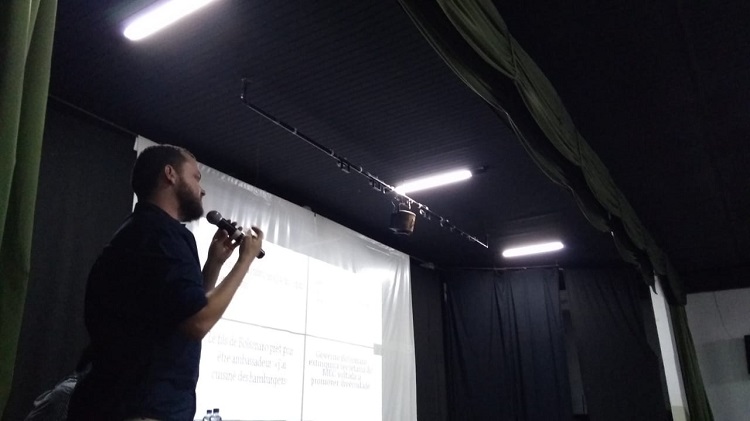 Palestrante, em pé, segurando o microfone, pele clara, camisa preta, barba